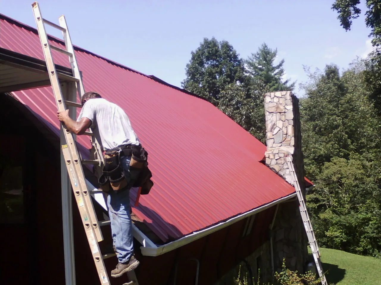 Roofer on a ladder beside a red metal roof and stone chimney — Soffit & Fascia Repair by Roofing Brothers in Johnstown